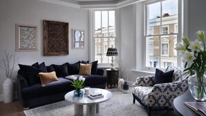 A modern living room with a dark blue sofa, a patterned armchair, and a round coffee table. There are framed artworks on the wall and a large window providing a view of buildings. - 317 Grand Maisonette (London)
