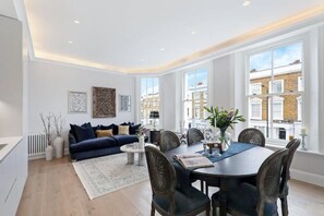 A modern living room with a dark blue sofa, a dining table with chairs, and large windows offering a view of buildings. - 317 Grand Maisonette (London)