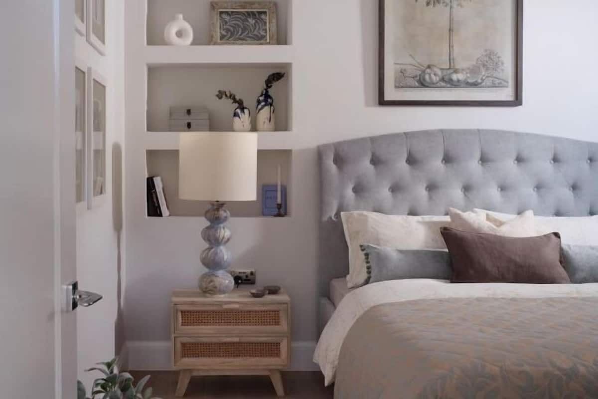A bedroom with a tufted headboard, a bedside table with a lamp, and a wall-mounted shelf with decorative items.