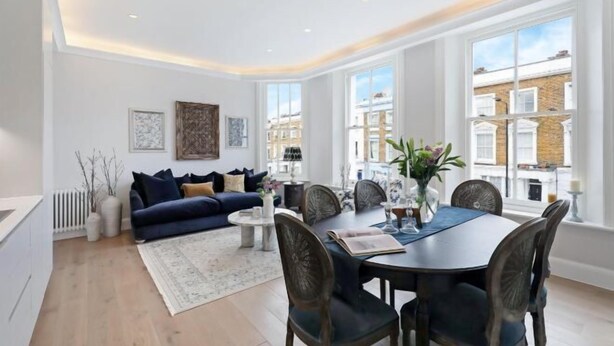 A modern living room with a dark blue sofa, a round dining table, and large windows offering a view of the city. - 317 Grand Maisonette (London)