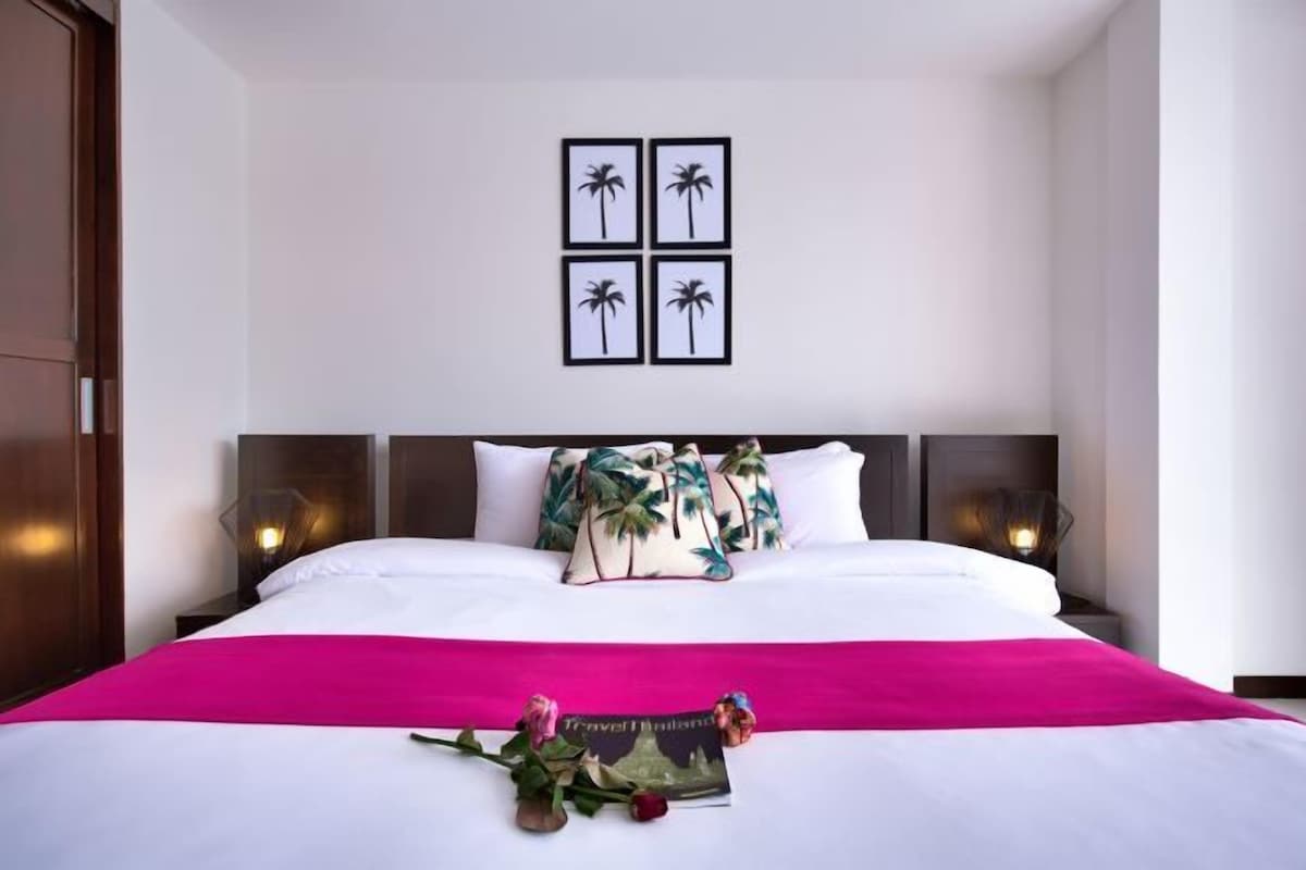 A neatly made bed with a white and pink bedspread, a decorative pillow with palm leaves, and a framed picture of palm trees on the wall.