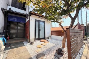 Un bâtiment avec une porte fermée, un petit espace extérieur agrémenté de pas japonais et un arbre qui offre de l’ombre.