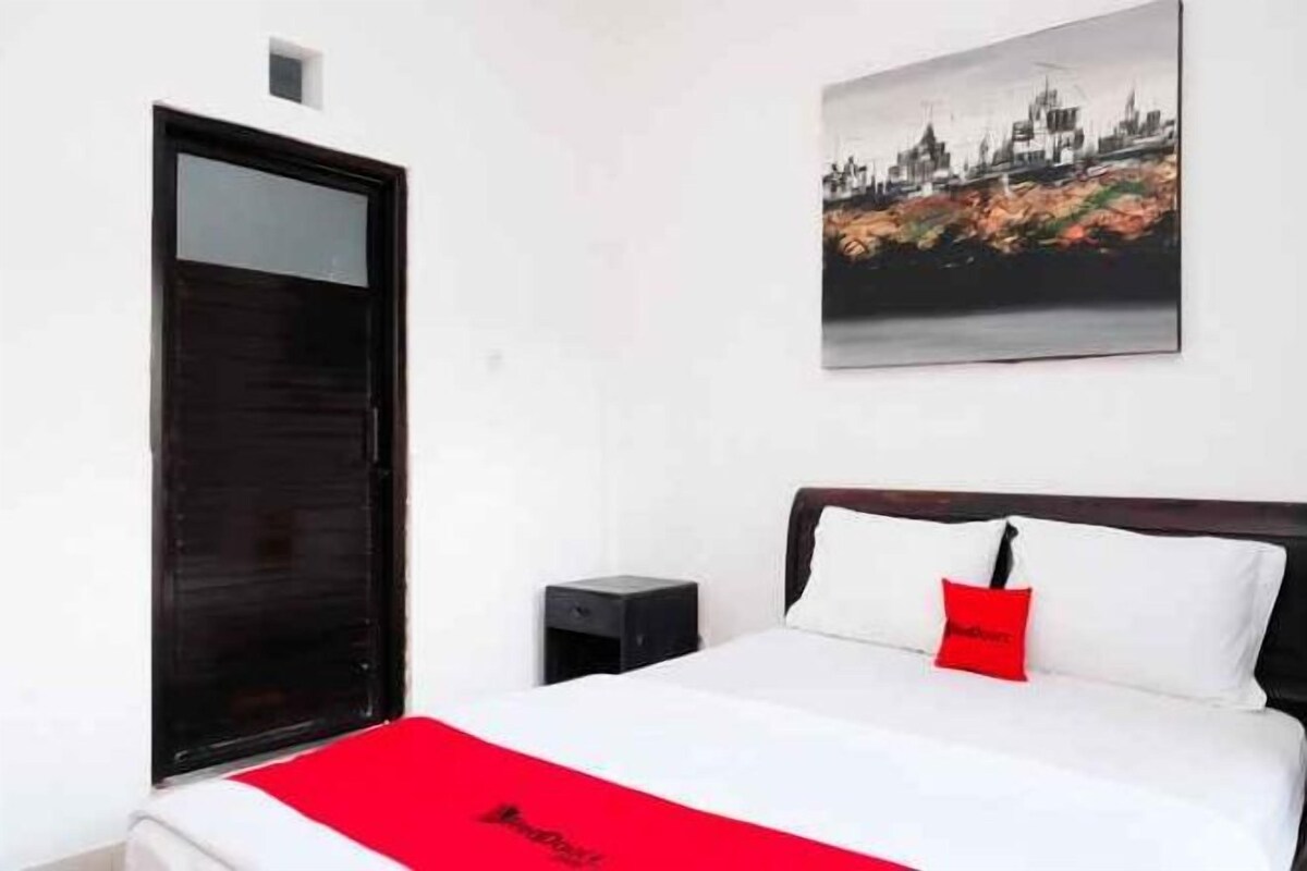 Une chambre d’hôtel avec un lit, un plaid rouge, un tableau représentant un paysage urbain, une porte noire et une petite table.