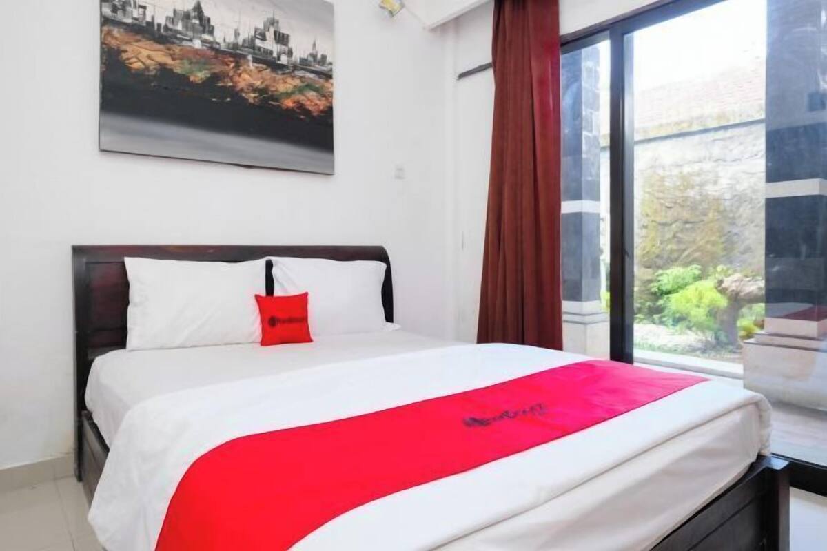 Une chambre d’hôtel avec un lit, une couverture rouge, un tableau représentant un paysage urbain et une fenêtre donnant sur un espace verdoyant.