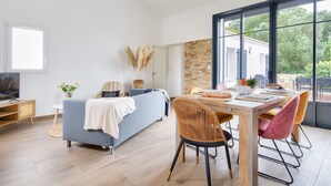 A modern living room with a sofa, dining table, and chairs, a TV, and a large window with a view of greenery. - Beach 400 m away - House for 4 with terrace (La Guérinière)