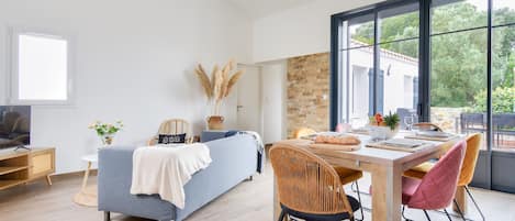 Ein modernes Wohnzimmer mit einer Couch, einem Esstisch mit Stühlen, einem Fernseher und einem großen Fenster mit Blick auf Grünflächen.