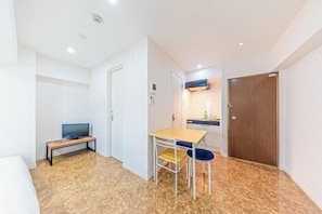 A compact room with a bed, a television on a stand, a small table with two chairs, a wooden door, and a kitchenette with a stove. - Motomachi Hisui Building (Yokohama)