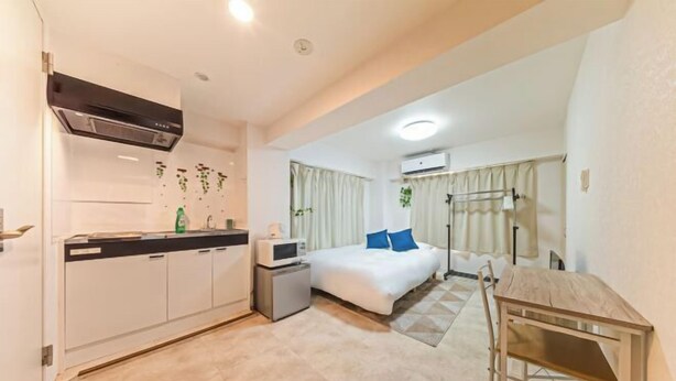 A compact hotel room with a kitchenette, a bed with blue pillows, a desk, and a window with curtains. - Motomachi Hisui Building (Yokohama)