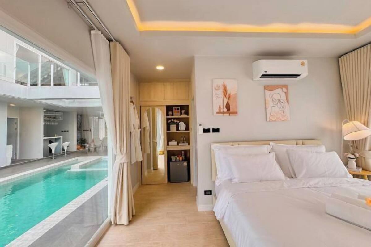A hotel room with a large bed, a bedside table, an air conditioning unit, a painting, and a view of an indoor swimming pool.