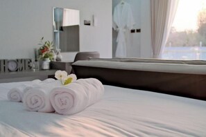 A neatly made bed with rolled towels and a flower, a bathroom with a mirror and towel rack, and a view of a building through the window. - The value of time pool villa Khaoyai (Pak Chong)