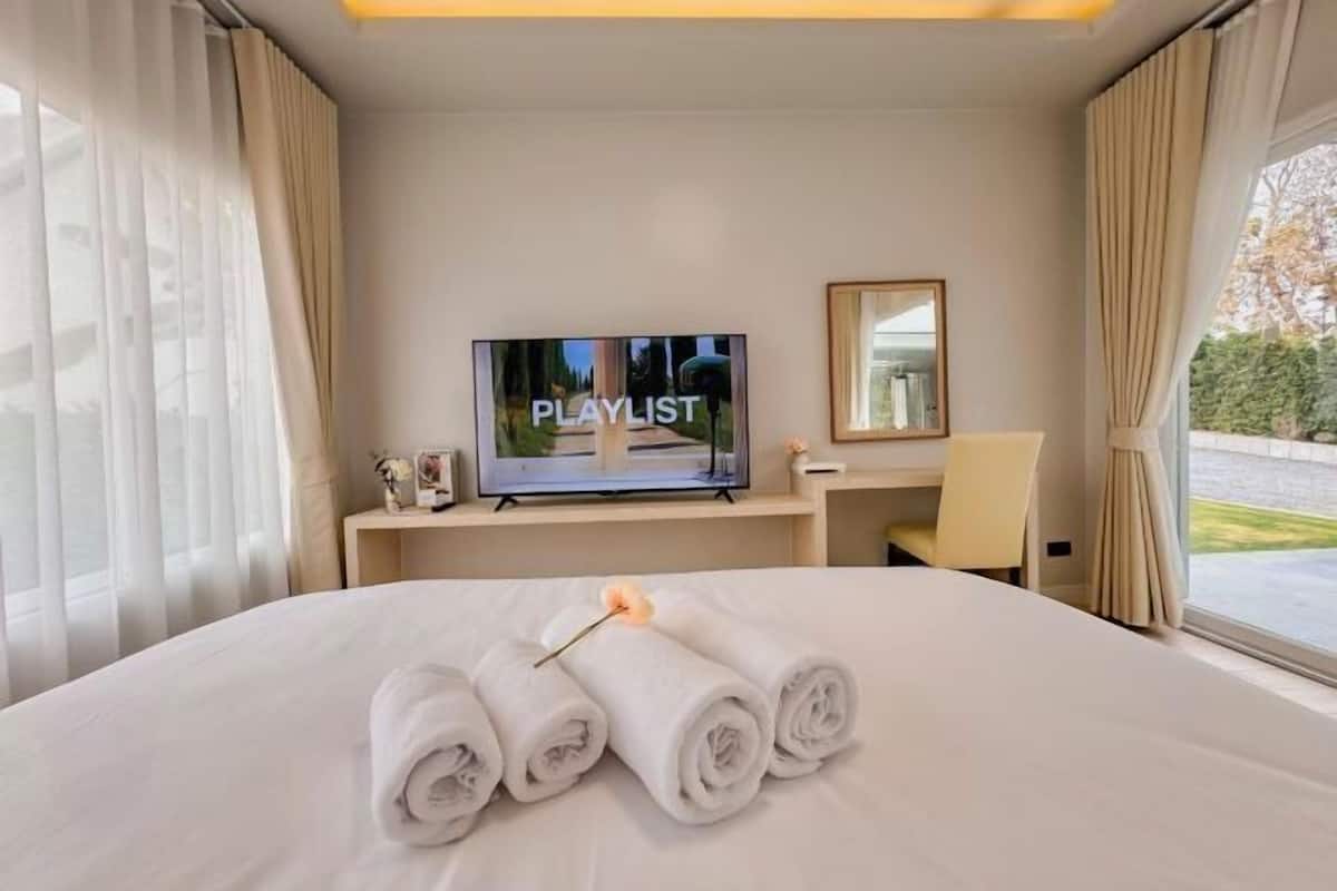 A hotel room with a bed, a television displaying 'PLAYLIST', a desk with a chair, and a window with curtains.