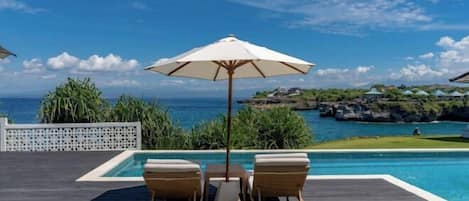 A poolside area with two lounge chairs and a table under a large umbrella, overlooking a clear blue sea and rocky coastline.