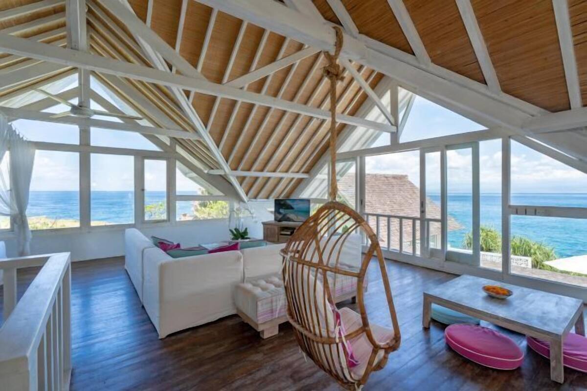 A room with a wooden ceiling, large windows, a hanging chair, a white sofa, and a small table.