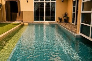 A swimming pool with a tiled edge, a grassy area, and a building with large windows. - Batu Pahat Santalia Villa 4BR 12pax Private Pool (Simpang Kanan)