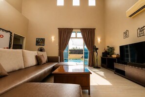 A living room with a sofa, coffee table, TV, and a view of a pool. - Batu Pahat Santalia Villa 4BR 12pax Private Pool (Simpang Kanan)