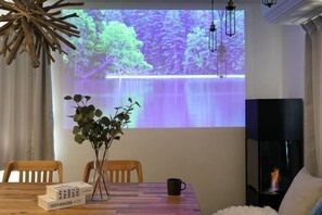 A room with a projector screen displaying a nature scene, a wooden dining table with chairs, a vase with green plants, and a fireplace.