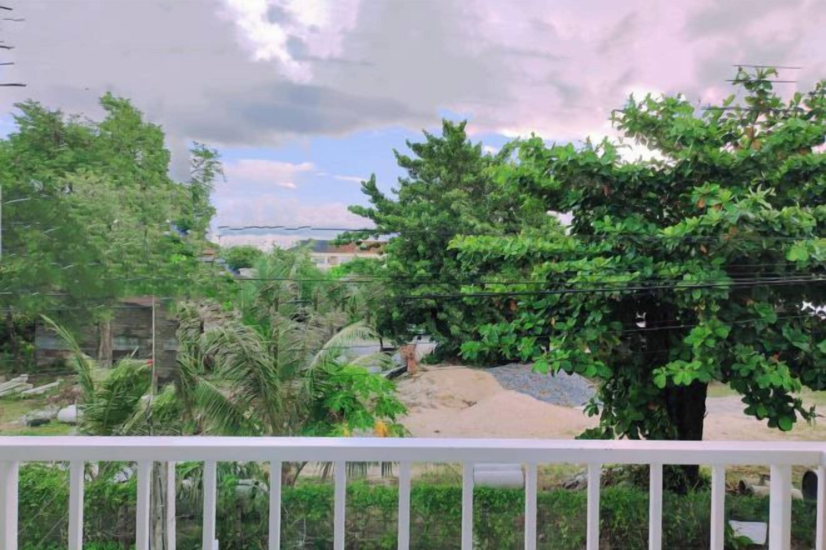 A balcony with a white railing, overlooking a garden with various trees and a clear sky.