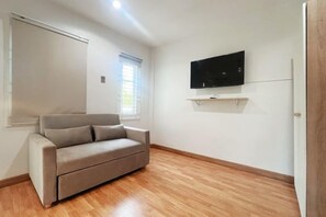 Ein Zimmer mit einer Couch, einem an der Wand montierten Flachbildfernseher und einem Fenster mit Jalousien.