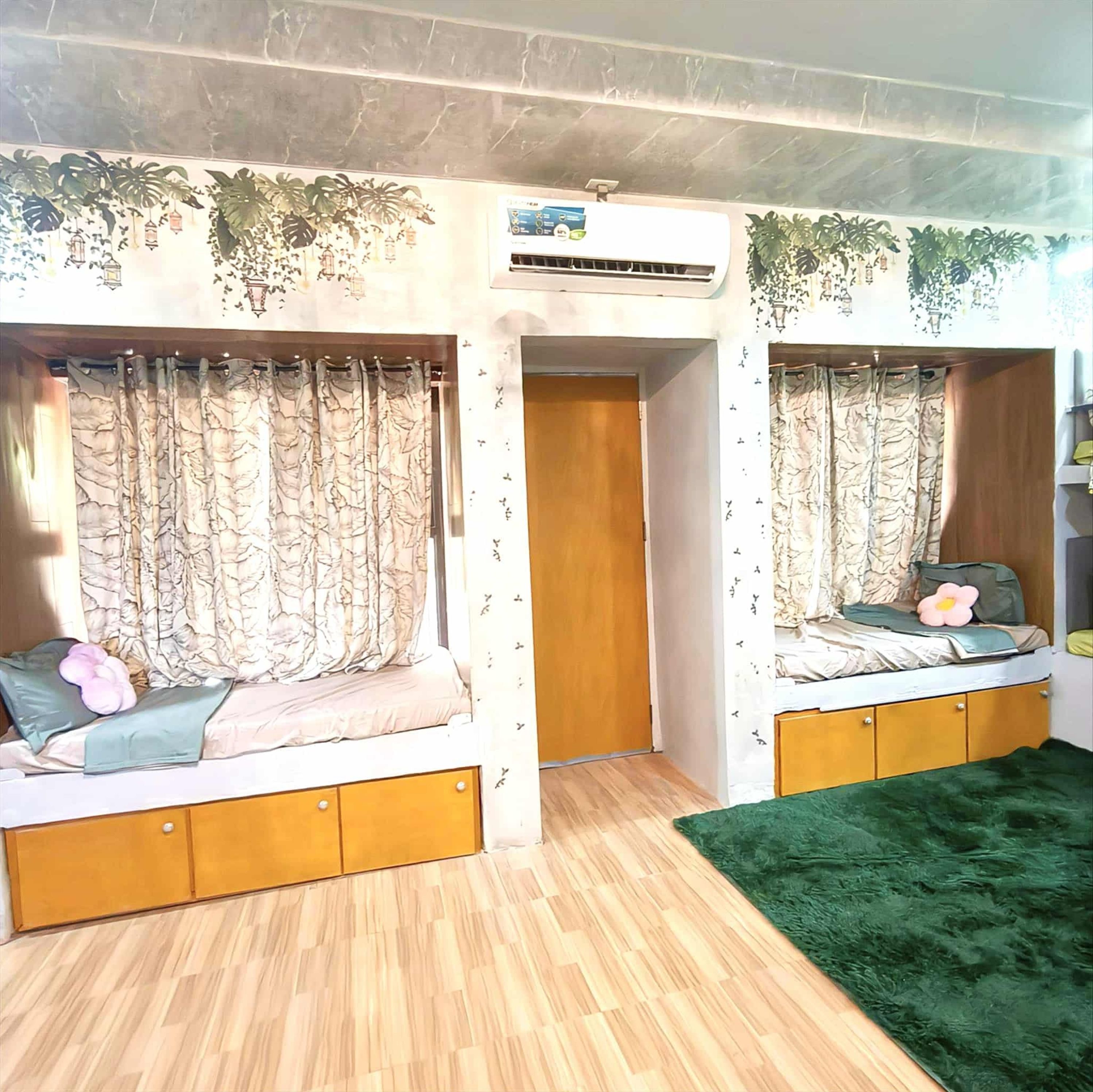 Une chambre avec deux lits superposés, chacun étant muni d’un rideau et d’un oreiller. On y trouve une porte en bois et un tapis vert sur le sol.