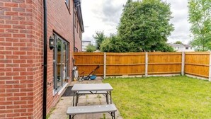 Een achtertuin met een stenen huis, een houten schutting, een tafel en een bank.