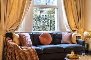 A room with a dark-colored sofa, a window with curtains, a decorative metal railing, and a small table with a lamp. - South Kensington - Cromwell Rd Residence (London)