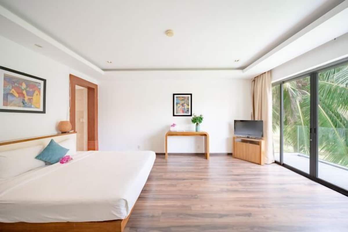 Een moderne hotelkamer met een groot bed, een televisie, een kleine tafel met een plant en een groot raam met uitzicht op palmbomen.