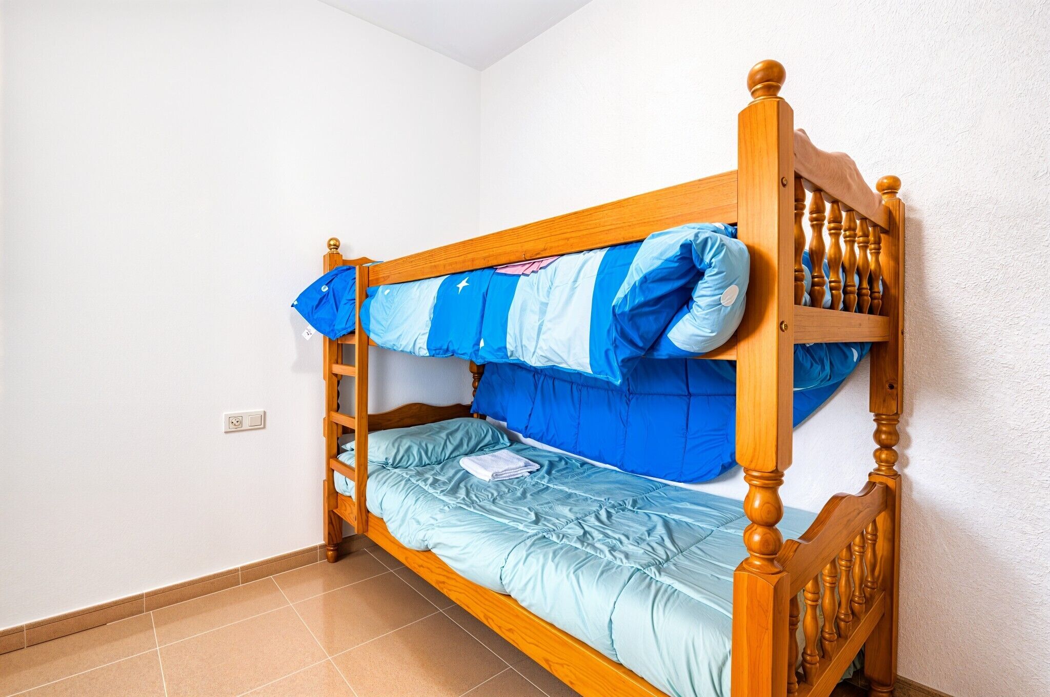 Un simple lit superposé en bois avec une échelle, une couverture à rayures bleues et blanches, et un matelas bleu clair.