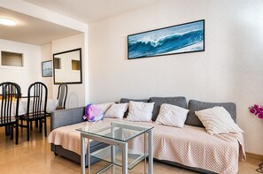 Una sala de estar con un sofá, una mesa de centro de vidrio, un comedor con sillas y un cuadro en la pared.