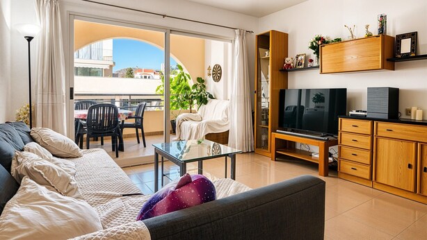 A living room with a sofa, a glass coffee table, a TV, a wooden cabinet, and a balcony with a dining area.