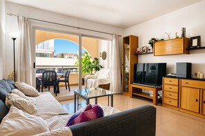 Ein Wohnzimmer mit einer Couch, einem Glastisch, einem Fernseher, einem Holzschrank und einem Balkon mit Essbereich.