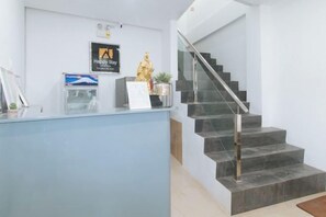 Una recepción con un cartel que dice 'Happy Stay', un cuadro enmarco y una pequeña estatua. Se ve una escalera con barandilla metálica que sube a un piso superior.