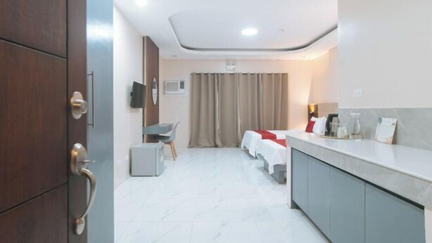 A hotel room with a bed, a desk, a chair, a small table, a TV, a window with curtains, and a kitchenette area. - RedDoorz @ Happy Stay Apartelle (Dauis)
