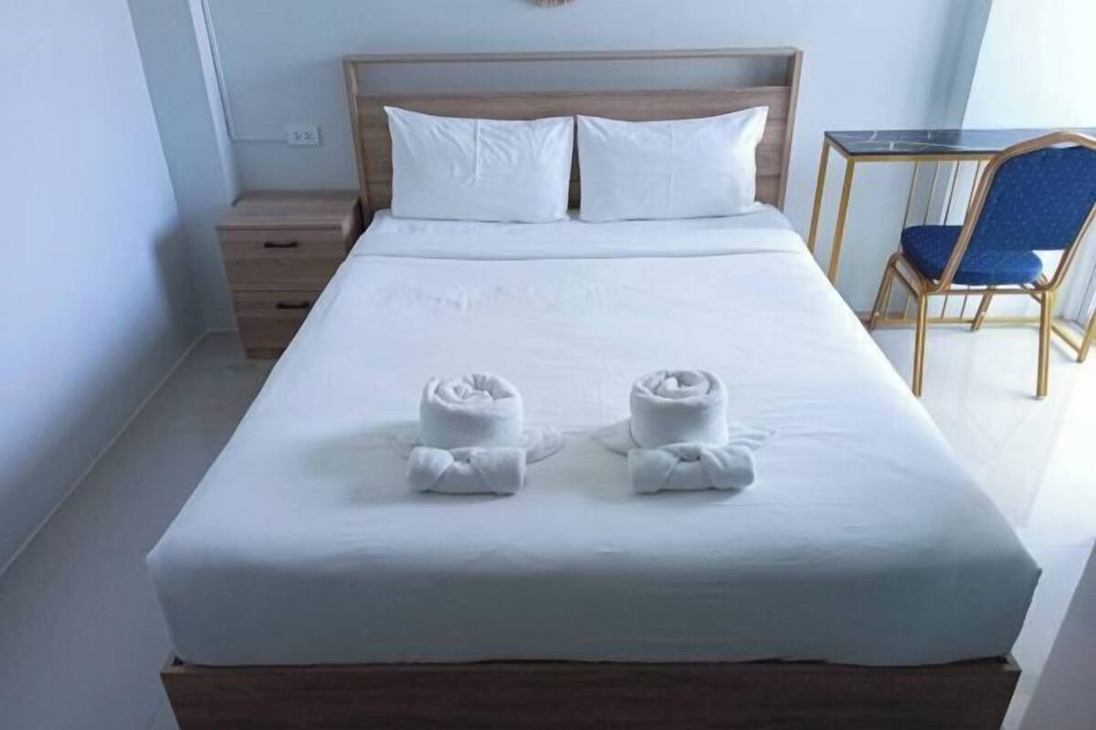 Un lit bien fait, avec deux serviettes enroulées, une tête de lit en bois, une petite table de chevet et une chaise avec un coussin bleu.
