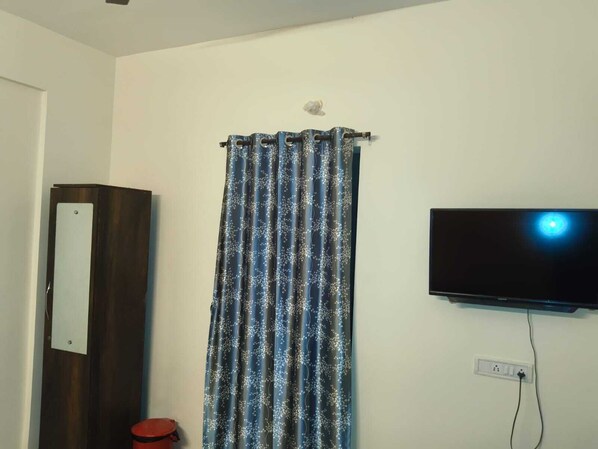 A room with a dark wooden wardrobe, a blue patterned curtain, a mounted TV, and a red stool.