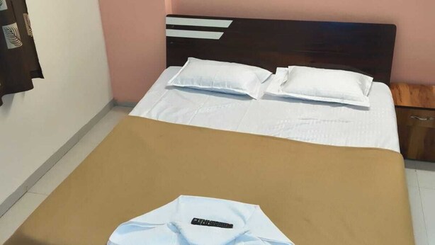 A neatly made bed with white bedding and a brown cover, a remote control on the bed, and a wooden nightstand beside it. - Balaji Lodging  (Hinjawadi)