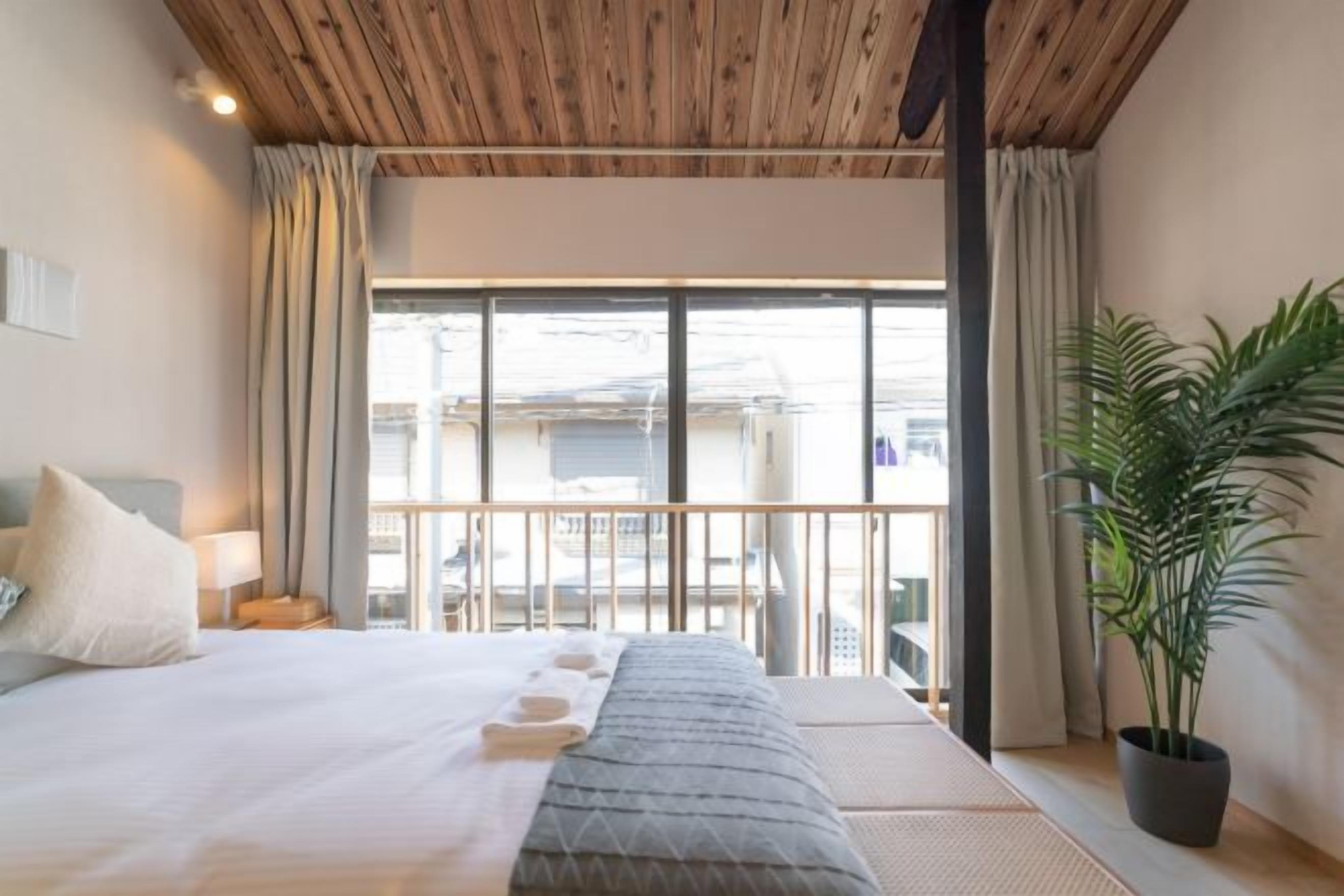 A hotel room with a bed, a potted plant, a balcony with a view, and a wooden ceiling.