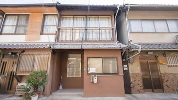 A two-story residential building with a traditional Japanese architectural style, featuring a tiled roof, wooden doors, and large windows with curtains.