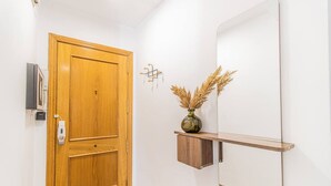 Un couloir avec une porte en bois, un miroir fixé au mur, une petite étagère sur laquelle se trouve un vase contenant des plantes séchées, et un dispositif de sécurité fixé au mur.