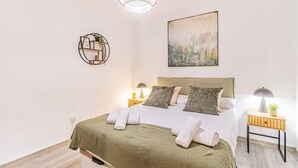 Un lit bien fait, avec une couverture verte et des serviettes blanches, deux lampes de chevet, une étagère fixée au mur avec une plante, et un tableau au-dessus du lit.