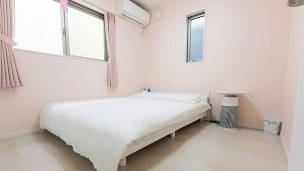 Quarto individual com uma cama branca, uma mesinha de cabeceira, um cesto de lixo e uma janela com cortinas.