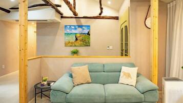 A room with a light blue sofa, wooden beams on the ceiling, a painting on the wall, and a small table with a plant.