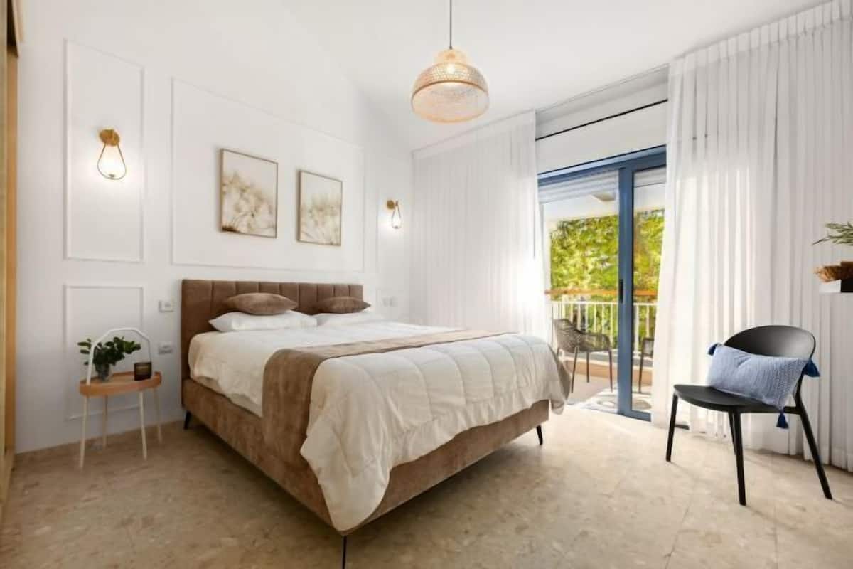 A modern bedroom with a large bed, a small side table, and a chair by the window.