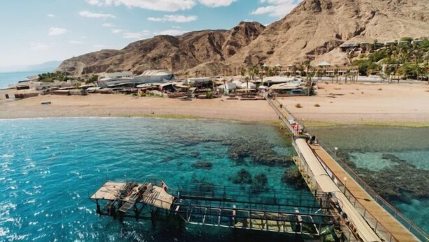 Une zone côtière avec une jetée s’avançant dans une eau d’un bleu limpide, un paysage montagneux en arrière-plan et une plage bordée de bâtiments et de palmiers.