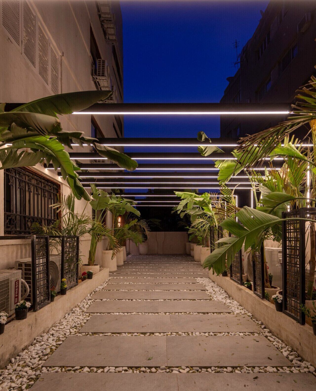 A pathway leading to a building entrance, flanked by potted plants and a modern light fixture.
