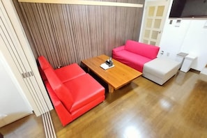 A room with a wooden table, a red sofa, a white ottoman, and a white cabinet. - Amistá.You (Osaka)