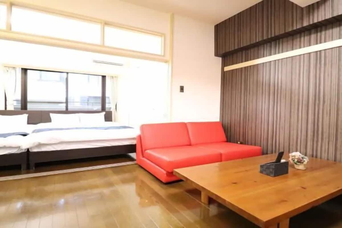 A room with a bed, a red sofa, a wooden table, and a window.