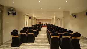 A hotel conference room with rows of black chairs with gold sashes, a podium at the front, and ceiling fans. - Kanak Residency Thalassary (Thalassery)