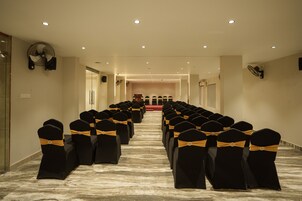 Un salón de conferencias de hotel con filas de sillas negras con cintas doradas, un atril al frente y ventiladores de techo.