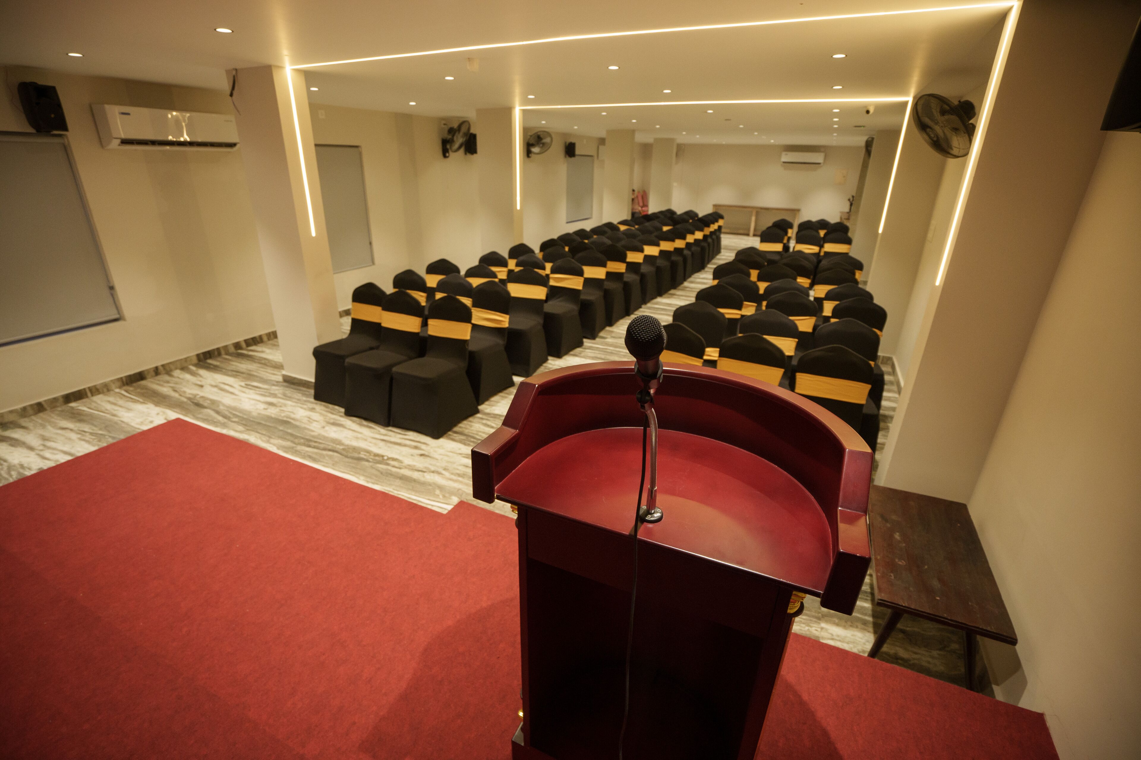 A conference room with rows of black chairs with yellow sashes, a red podium, and a marble floor.