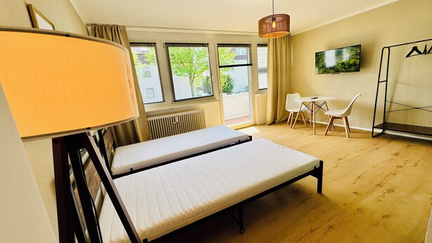 A room with a bed, a table, chairs, a TV, and a window with a view of trees. - Apartment Sana Klinik No 12 Balcony (Offenbach am Main)
